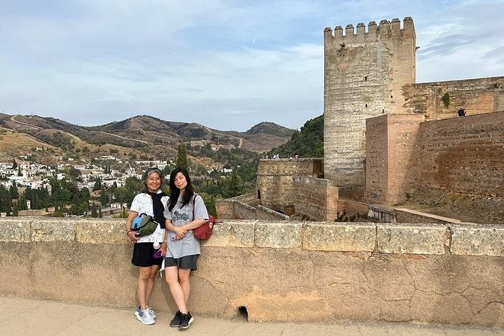 Two visitors beside an Alhambra stone wall and square tower, with Granada behind